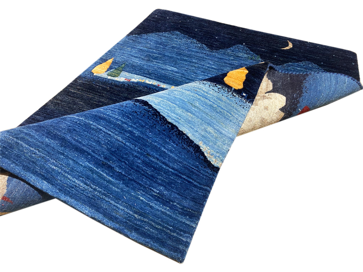 Gabbeh-Luri Teppich mit diagonaler Ansicht: Blau mit Berglandschaft und beigen Hausformen.