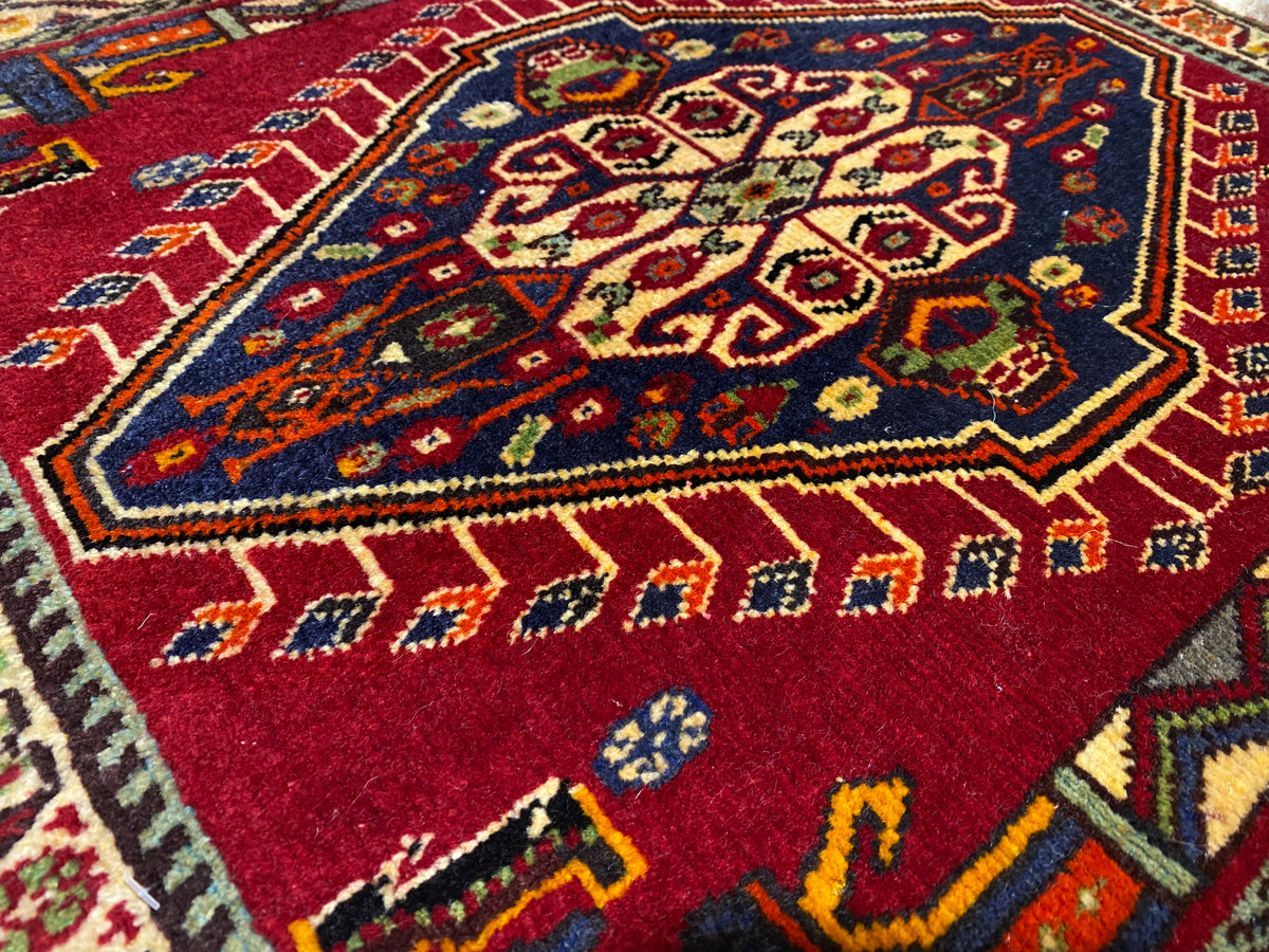 Primo piano di un tappeto Poshti (59×61 cm): motivo interno rosso con ornamenti colorati e bordo